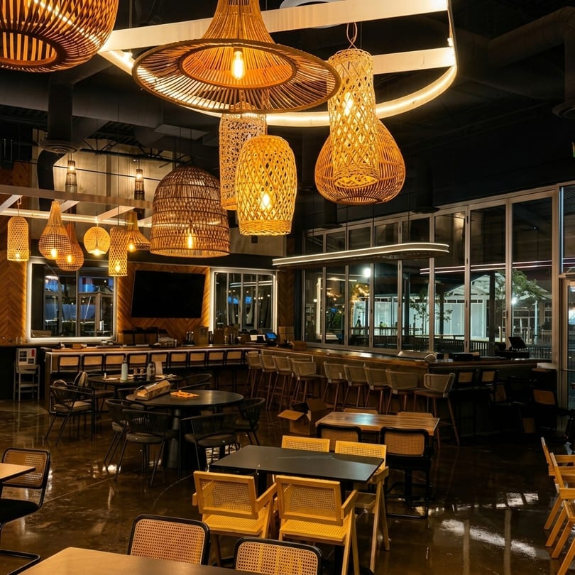 Restaurant interior with wicker light fixtures, a long bar, and dining tables with yellow and black chairs.