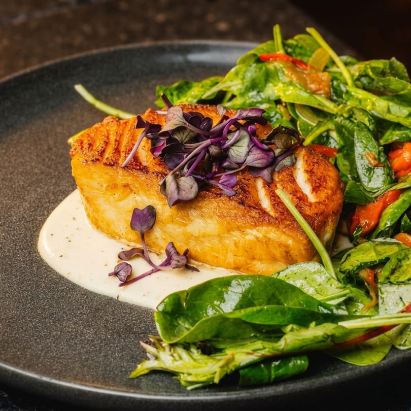 Pan-seared fish with purple microgreens, creamy sauce, and a side of fresh spinach salad.