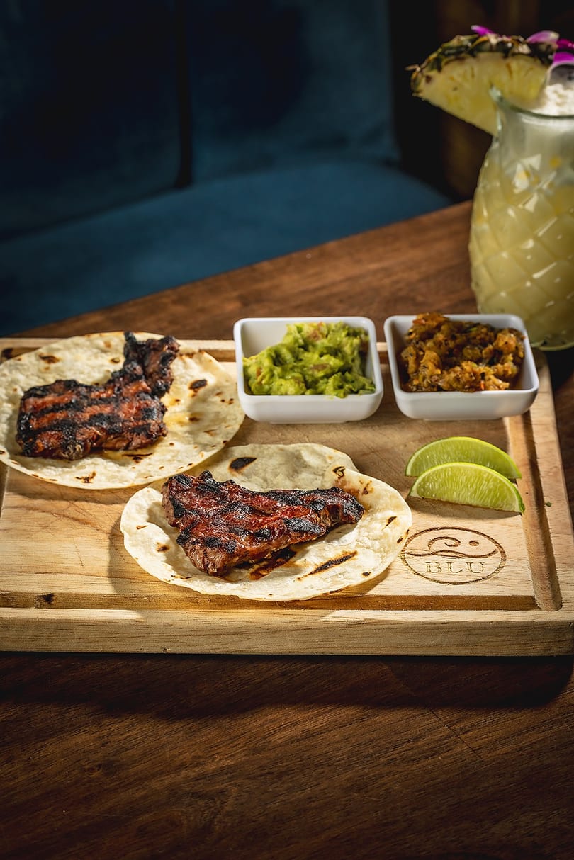 Grilled steak tacos with guacamole, salsa, and lime wedges on a wooden board with a pineapple drink.