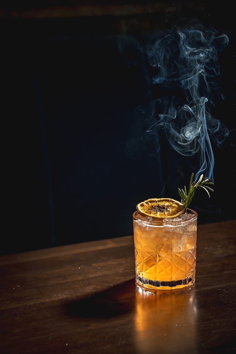 Smoked cocktail with a dried orange slice and rosemary sprig on a dark wooden bar.