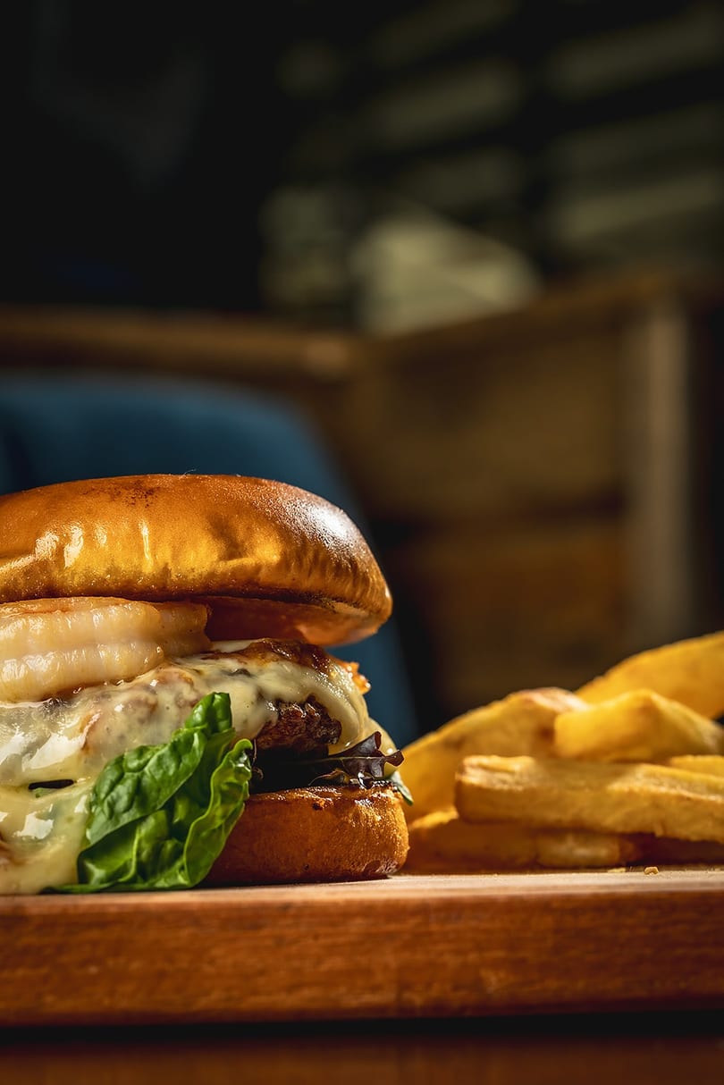 Gourmet burger with melted cheese, lettuce, and onion rings, served with fries on a wooden board.
