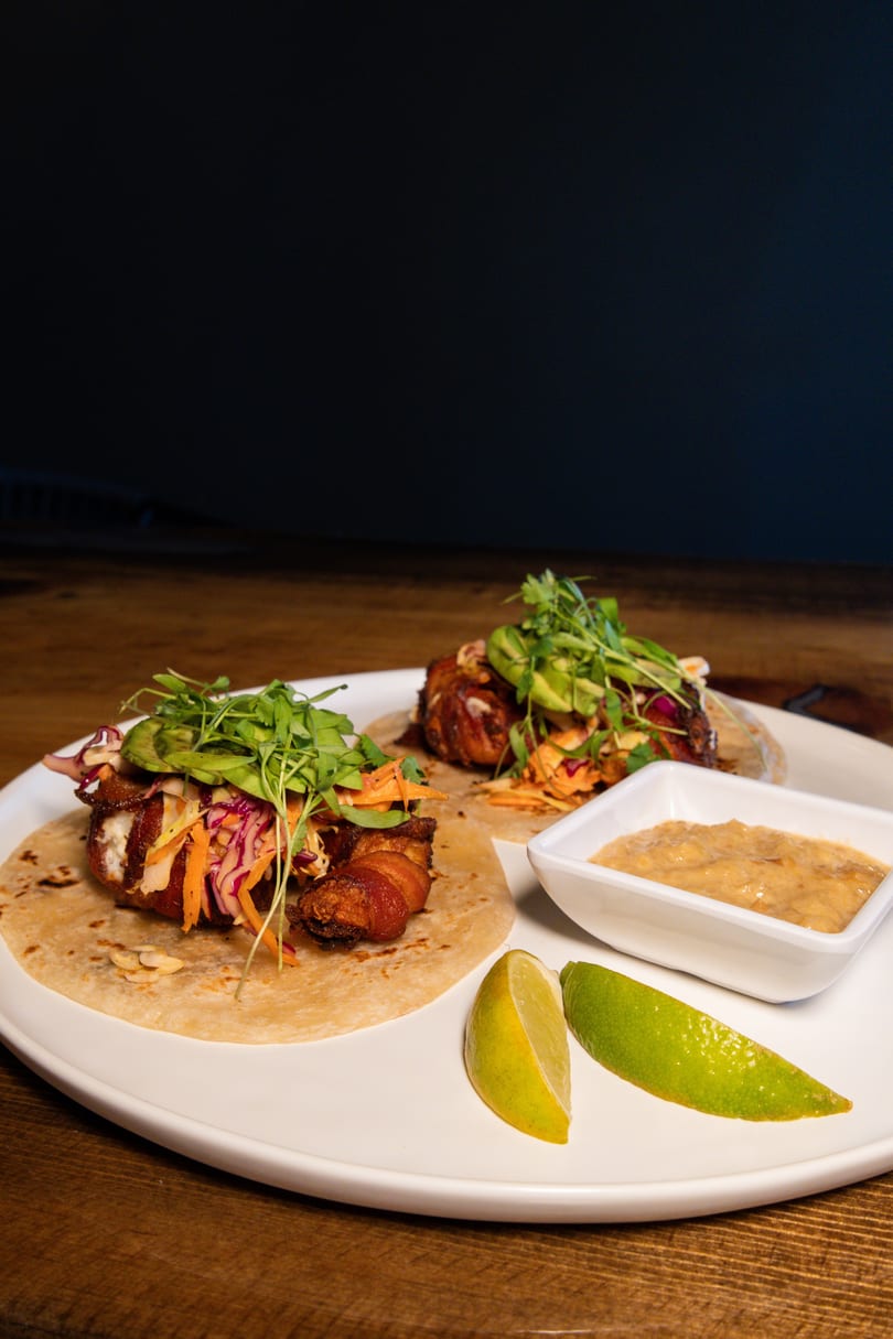 Two bacon-wrapped tacos with slaw, avocado, and microgreens, served with lime wedges and sauce.