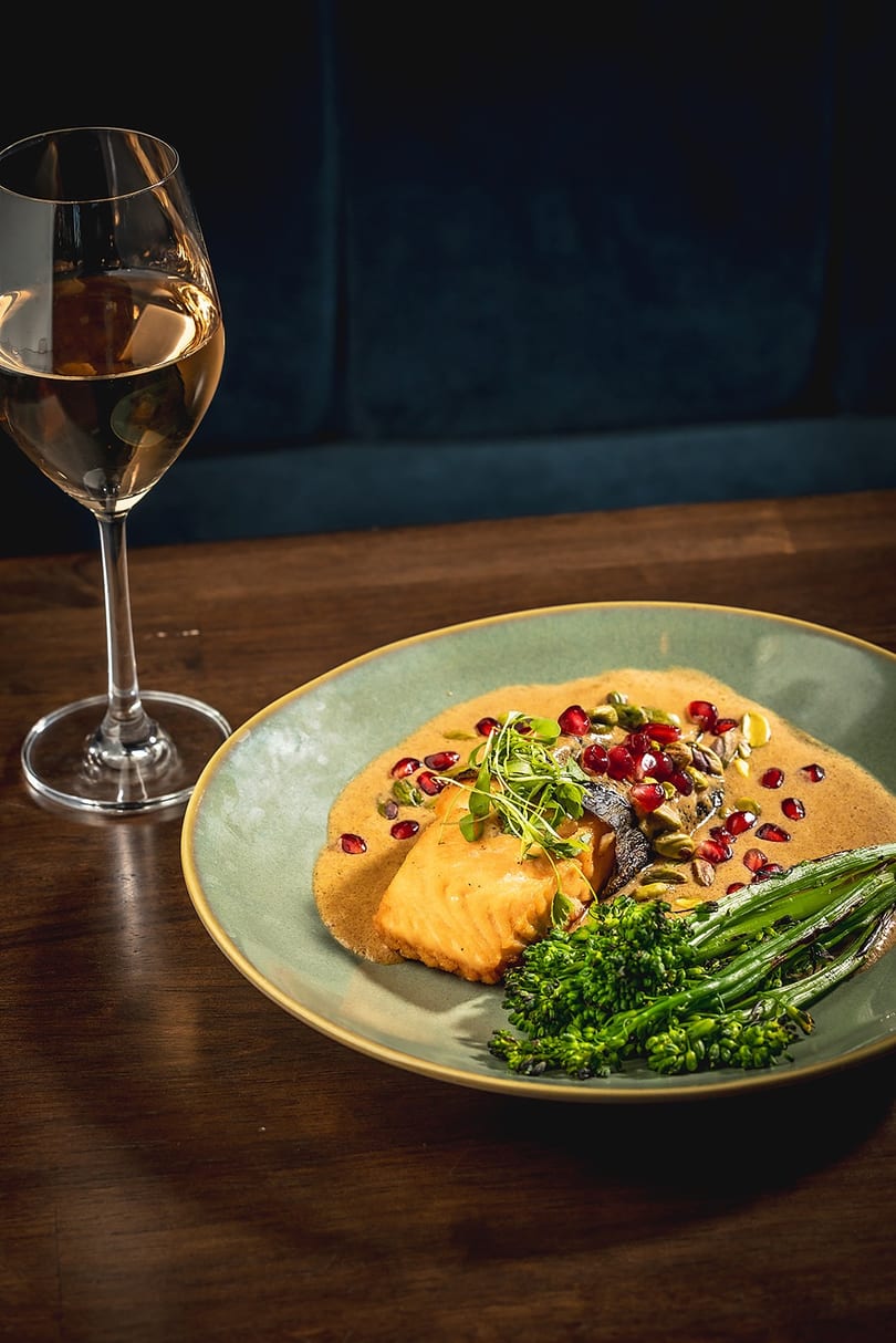 Salmon with mole sauce, pomegranate, pistachios, and broccolini, next to a glass of white wine.