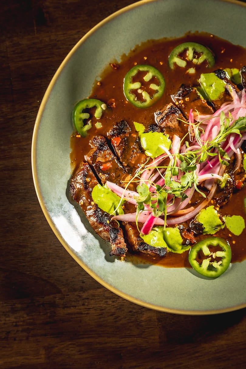 Grilled steak slices in a rich sauce, topped with pickled red onions, jalapeños, and green crema.
