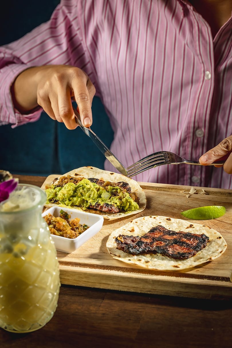 Person in a pink shirt cutting a taco with guacamole and grilled meat on a wooden board.