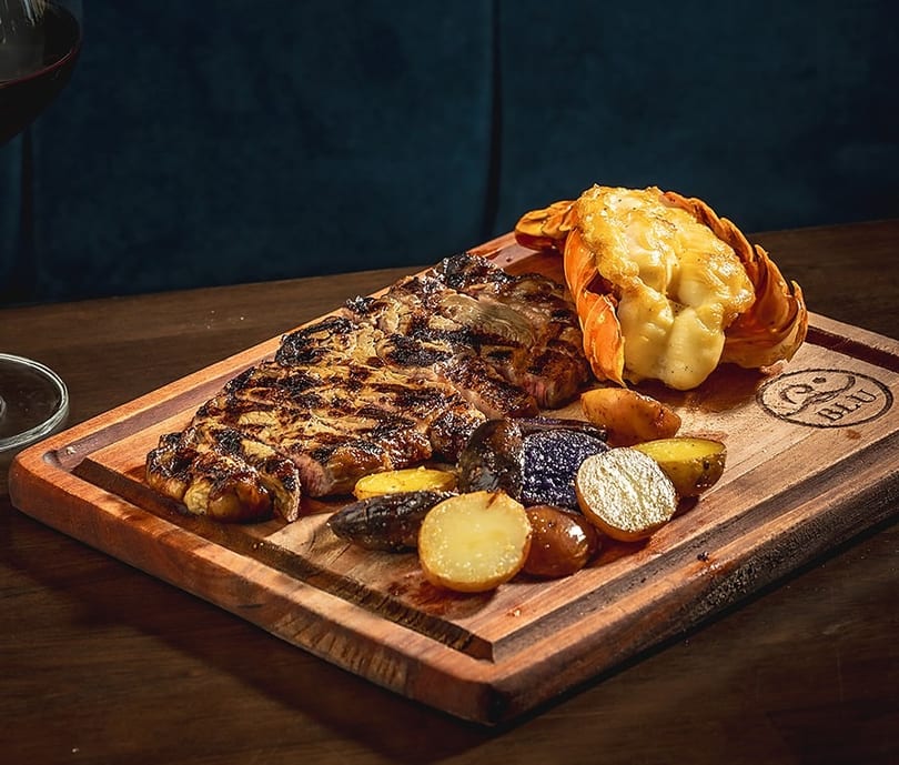 Grilled steak and lobster tail with roasted potatoes on a wooden board, with a glass of red wine.