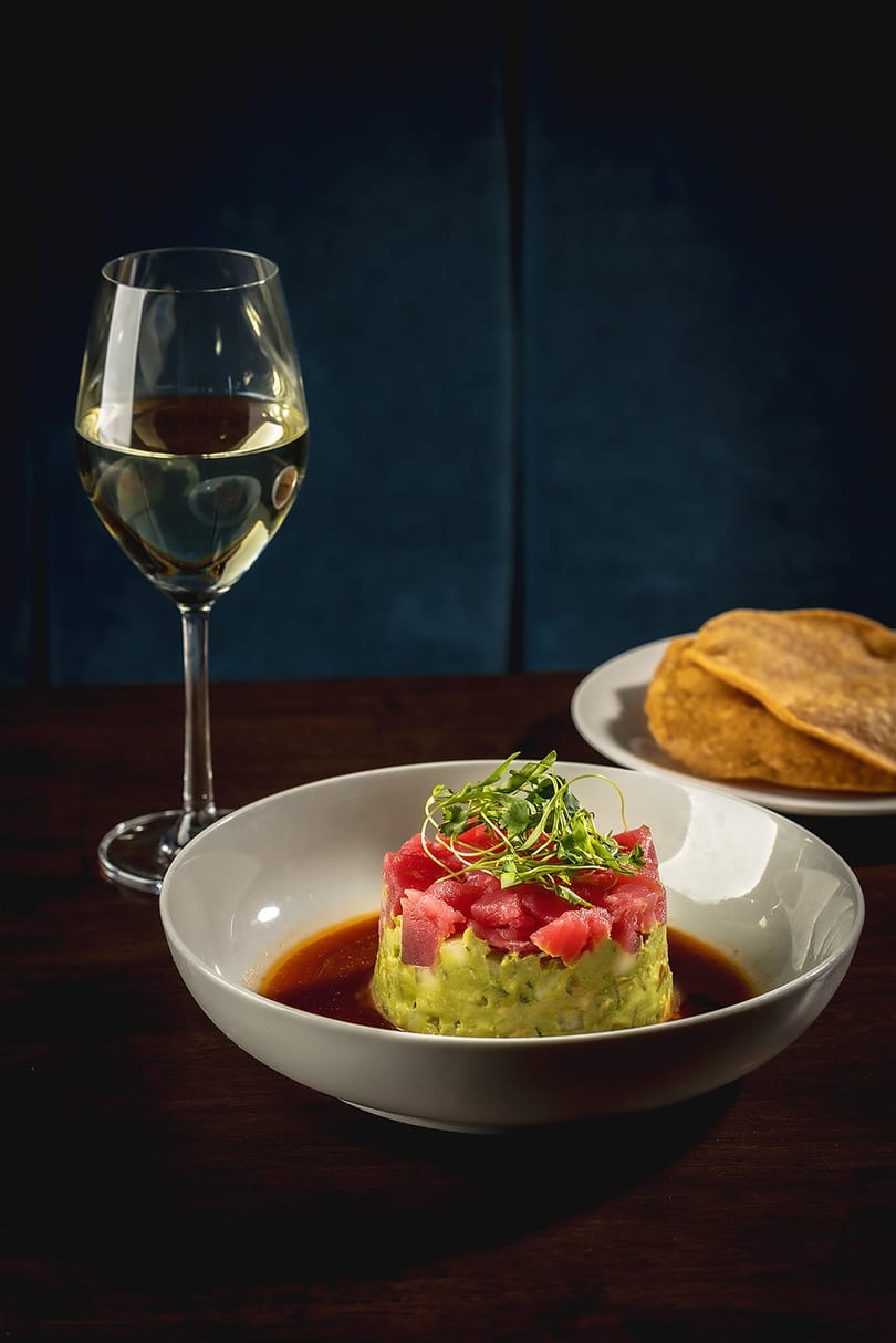 Tuna tartare with avocado, microgreens, and sauce, alongside a glass of white wine and tortillas.