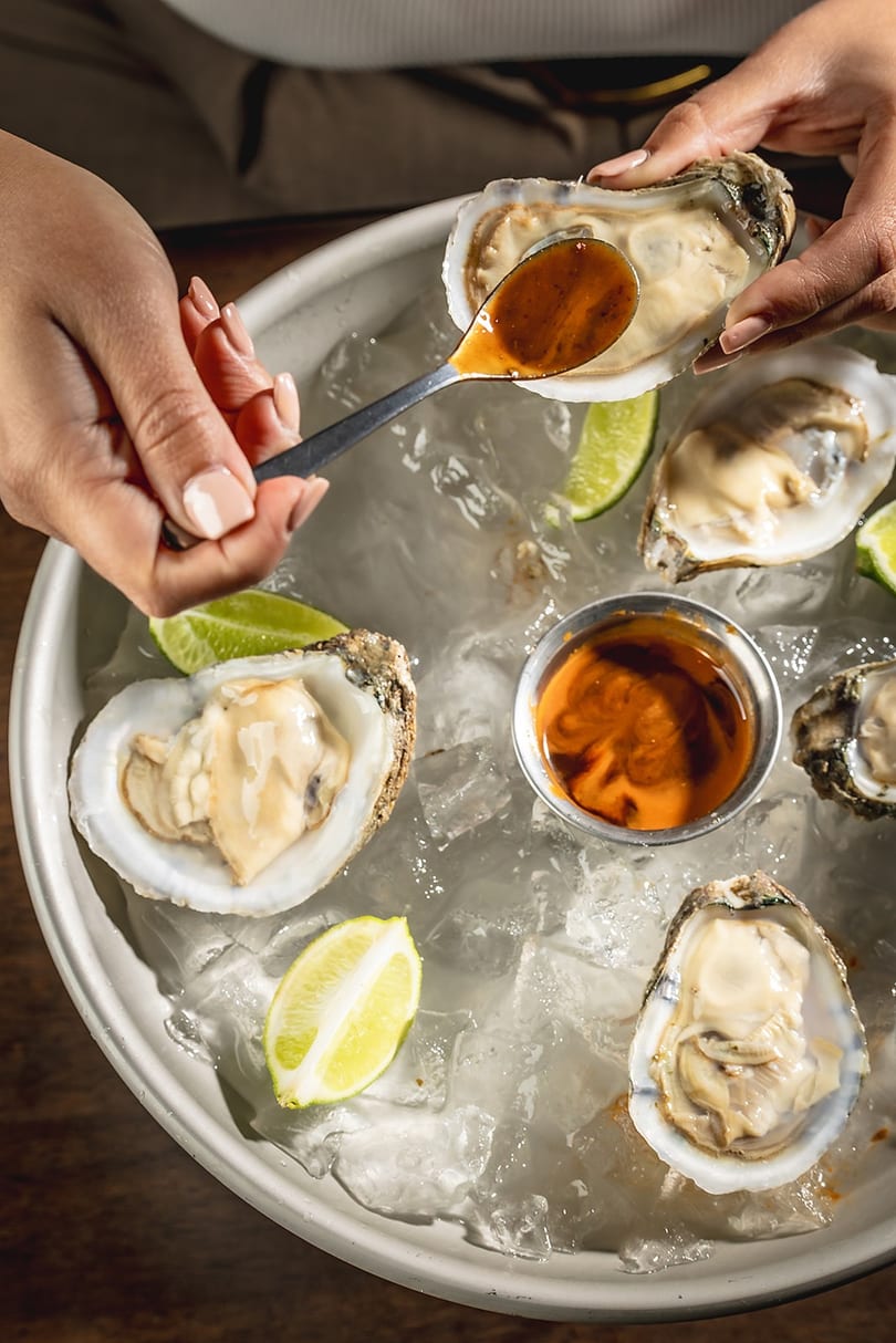 Hands spooning sauce onto a fresh oyster on a bed of ice with lime wedges.