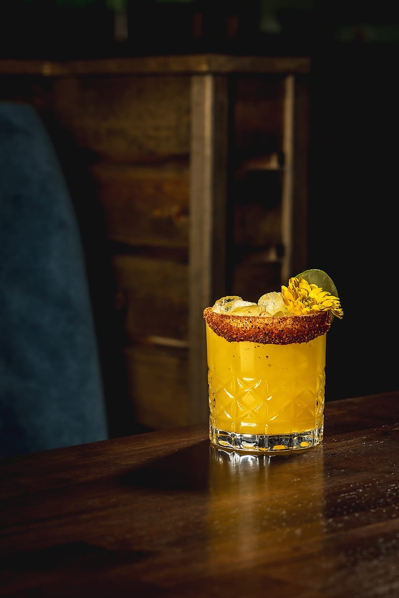 Yellow cocktail with a spiced rim and a yellow flower garnish on a dark wooden bar.