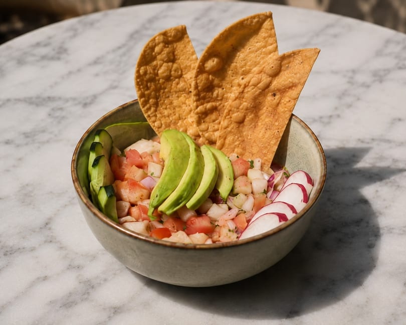 Ceviche bowl with avocado, cucumber, radish, and tortilla chips on a marble table.