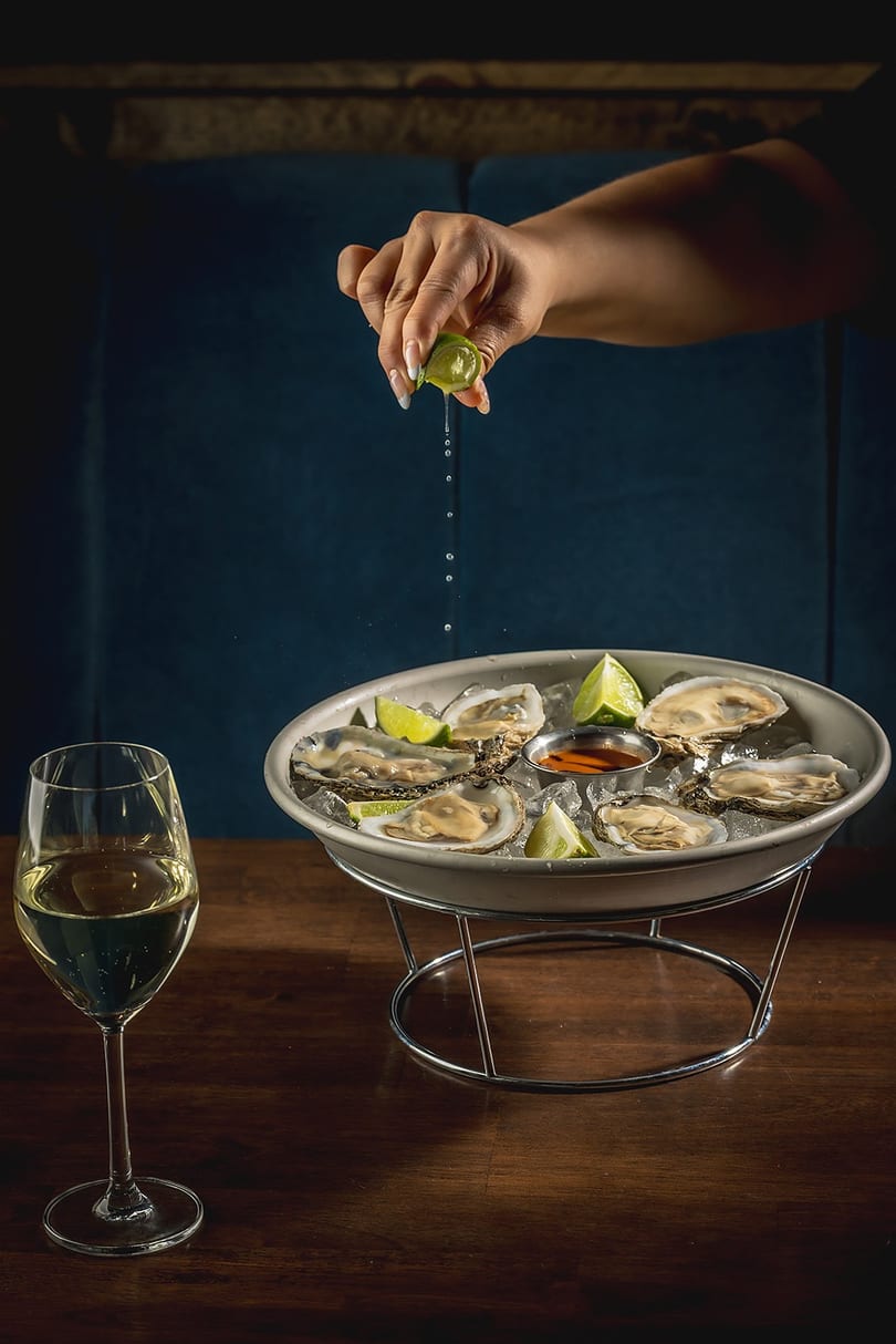 Hand squeezing lime over oysters on ice with a glass of white wine on a wooden table.