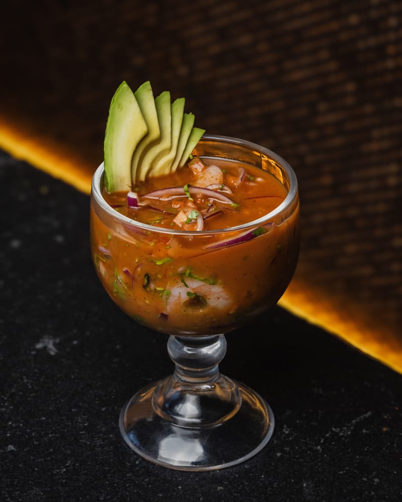 Shrimp ceviche in a glass goblet, garnished with avocado slices and red onion, on a dark surface.