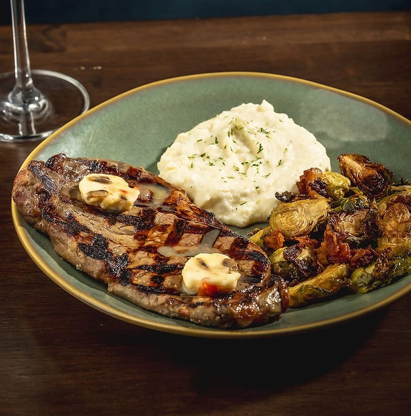 Grilled steak with melting butter, mashed potatoes, and roasted Brussels sprouts on a green plate.