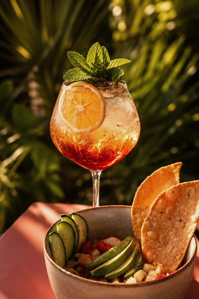 A vibrant orange cocktail with mint and citrus, next to a bowl of ceviche with tortilla chips.
