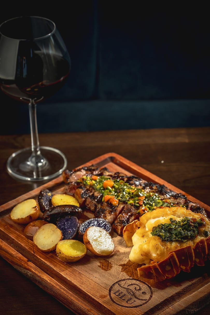 Grilled steak with chimichurri, roasted potatoes, and lobster tail on a wooden board with red wine.